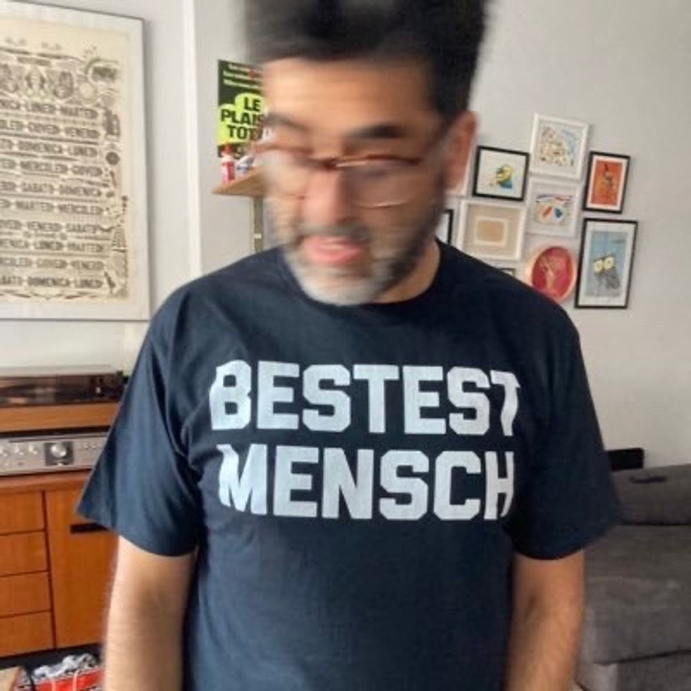 A person wearing a dark shirt with white text that reads "BESTEST MENSCH" standing in a room with a blurred background showing shelves, framed art, and furniture.